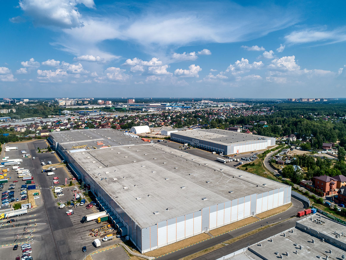 Huge warehouse building with flat roof and cars on parking site under blue sky with fluffy clouds near city in summer bird eye view