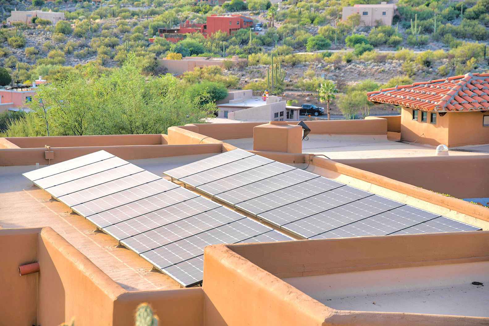 Solar panels on a flat roof of a mediterranean house at Tucson, Arizona. From the roof there is a view of a neighborhood with cactuses outdoors.