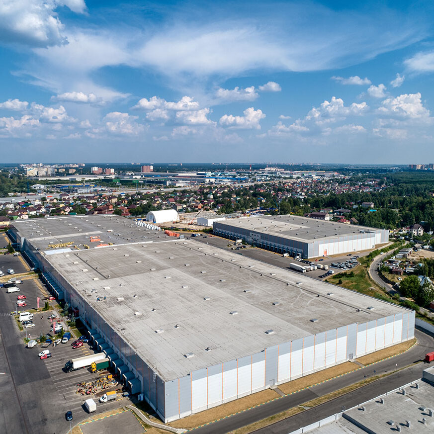 Huge warehouse building with flat roof and cars on parking site under blue sky with fluffy clouds near city in summer bird eye view